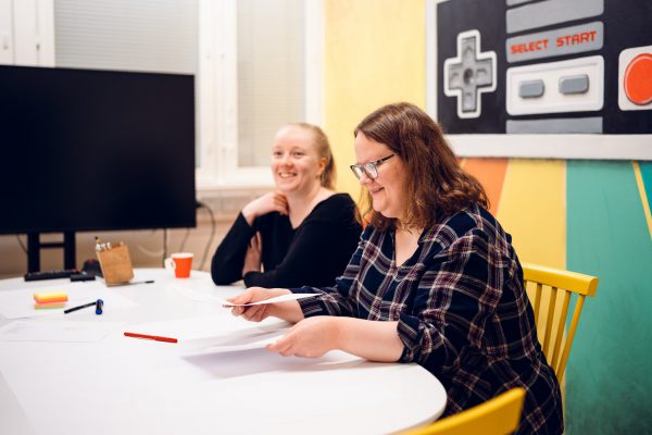 Kaksi naista istuu pöydän ääressä ja hymyilevät. Kameraa lähempänä olevalla naisella on paperia käsissään.
